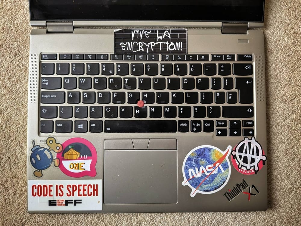 A top-down photo of the keyboard part of a silver-coloured laptop with black keys. A 'ThinkPad X1' symbol is visible along with various stickers.