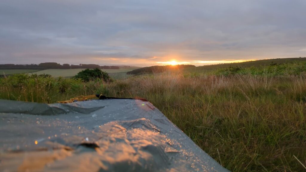 Part of a rain-covered tent with sunrise over trees in the distance.
