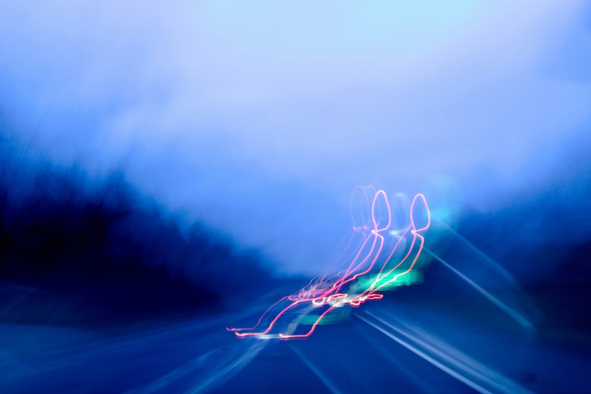 A blurry photo of a car driving down a highway. The red lights have made weird shapes, which could be interpreted in different ways.