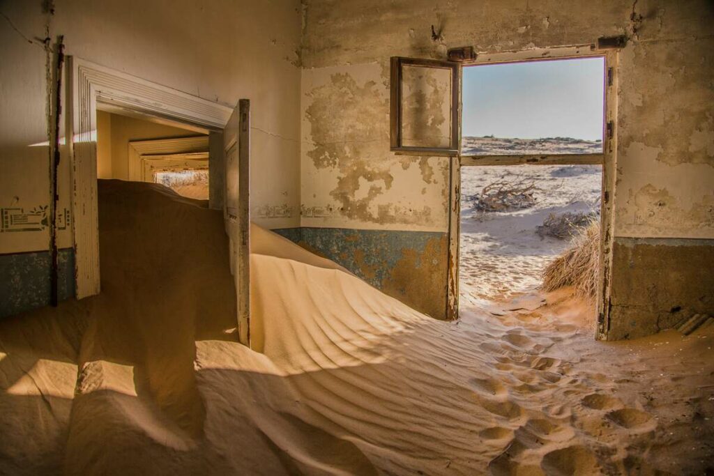 Abandoned building with sand filling rooms and desert landscape visible through an open door.

