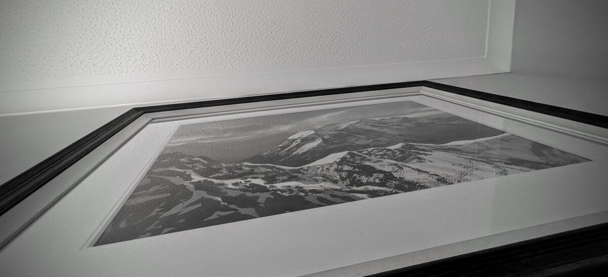 Looking upwards at a narrow angle at a painting of snowy mountains in a black frame.  