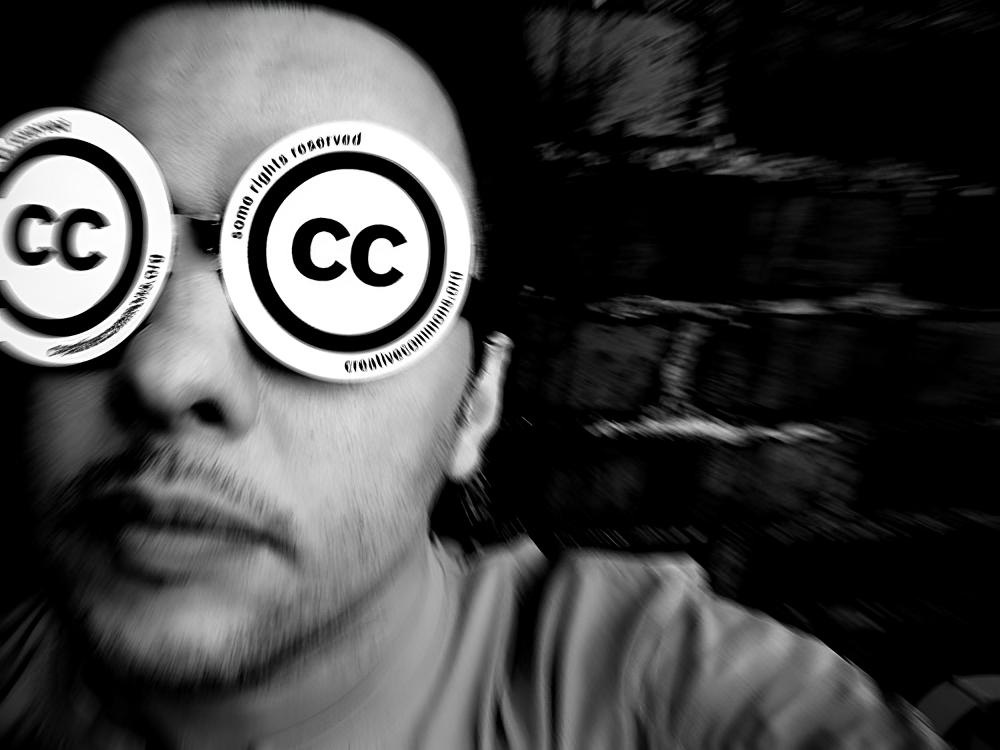 A slightly blurry black and white photo of a person wearing Creative Commons stickers over their sunglasses